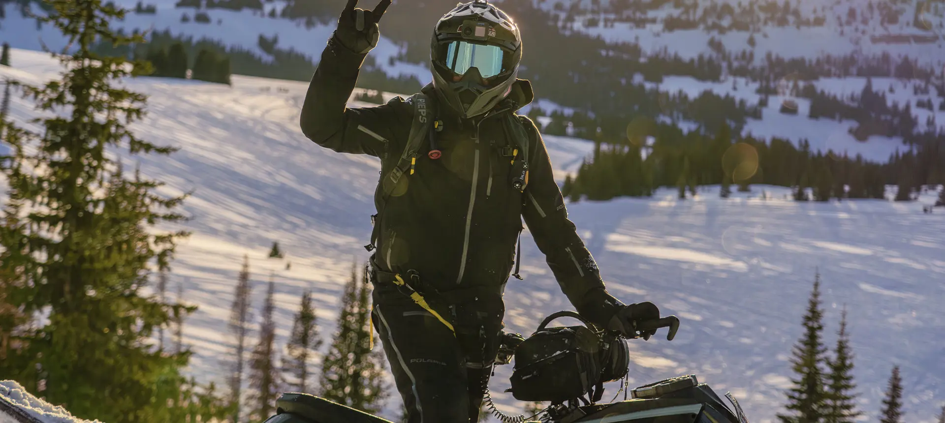 Helmeted Kyle Pulsifer signaling beside a snowmobile in a sunlit mountain landscape.