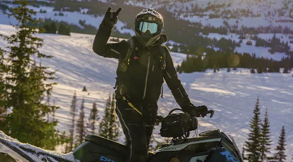 Helmeted Kyle Pulsifer signaling beside a snowmobile in a sunlit mountain landscape.