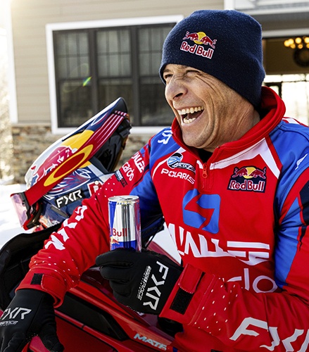 Levi LaValle in Red Bull racing gear holding a drink beside a red snowmobile in a snowy driveway.