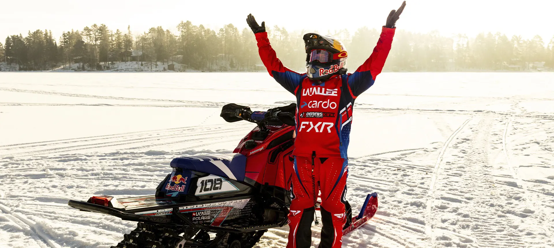 Levi LaValle raising both arms beside a custom red Polaris snowmobile on a frozen lake at sunset.