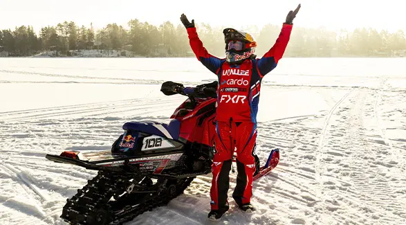 Levi LaValle raising both arms beside a custom red Polaris snowmobile on a frozen lake at sunset.