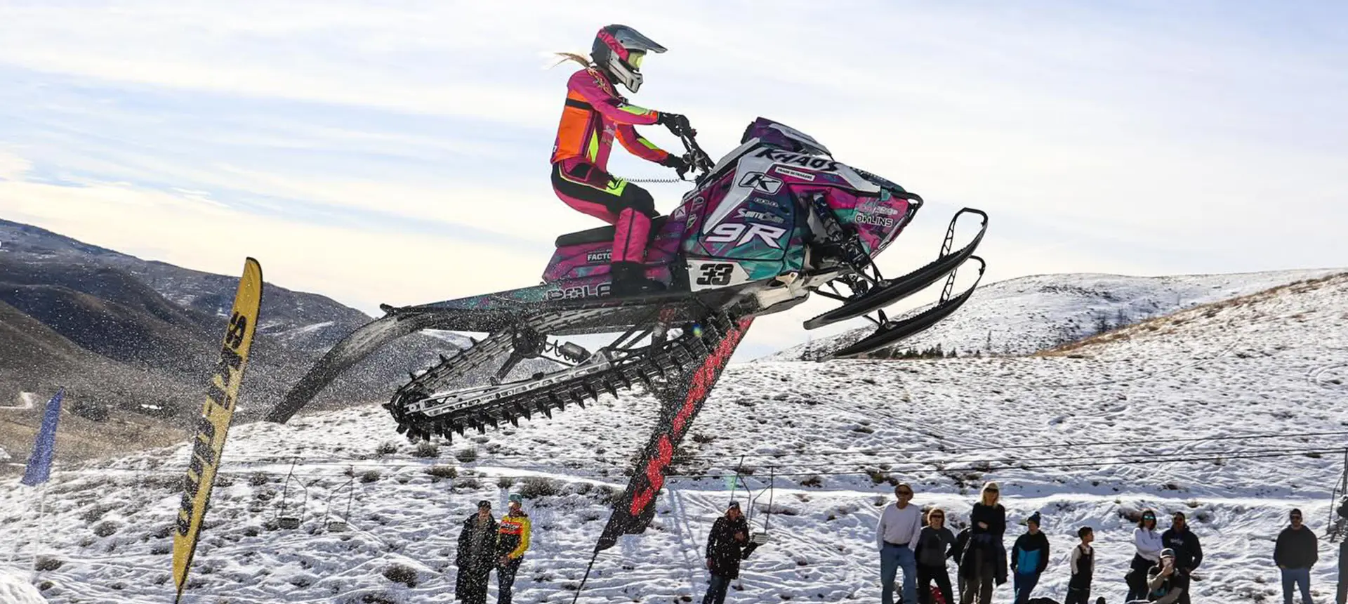 Mikaela Grönlund jumping a custom Polaris RMK Khaos snowmobile above spectators on a snowy hillside.