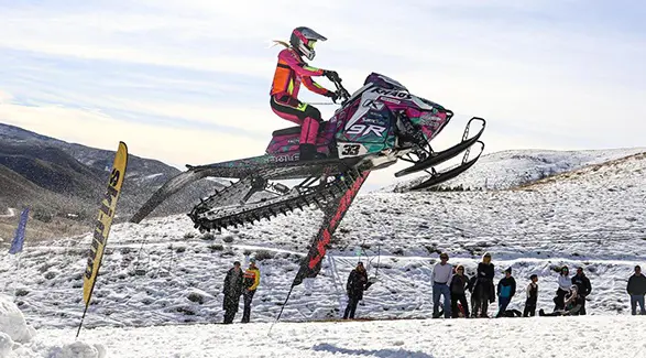 Mikaela Grönlund jumping a custom Polaris RMK Khaos snowmobile above spectators on a snowy hillside.