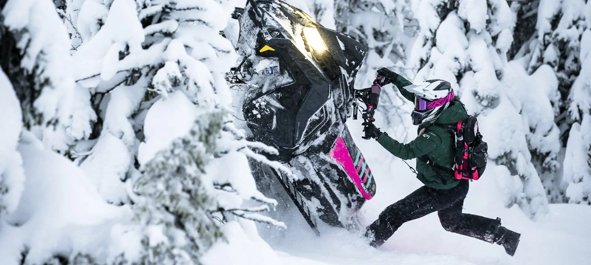 Mikaela Grönlund maneuvering a black and pink Polaris snowmobile through deep powder in a snow-covered forest.