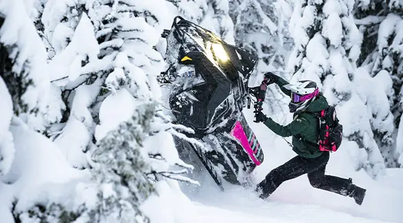 Mikaela Grönlund maneuvering a black and pink Polaris snowmobile through deep powder in a snow-covered forest.