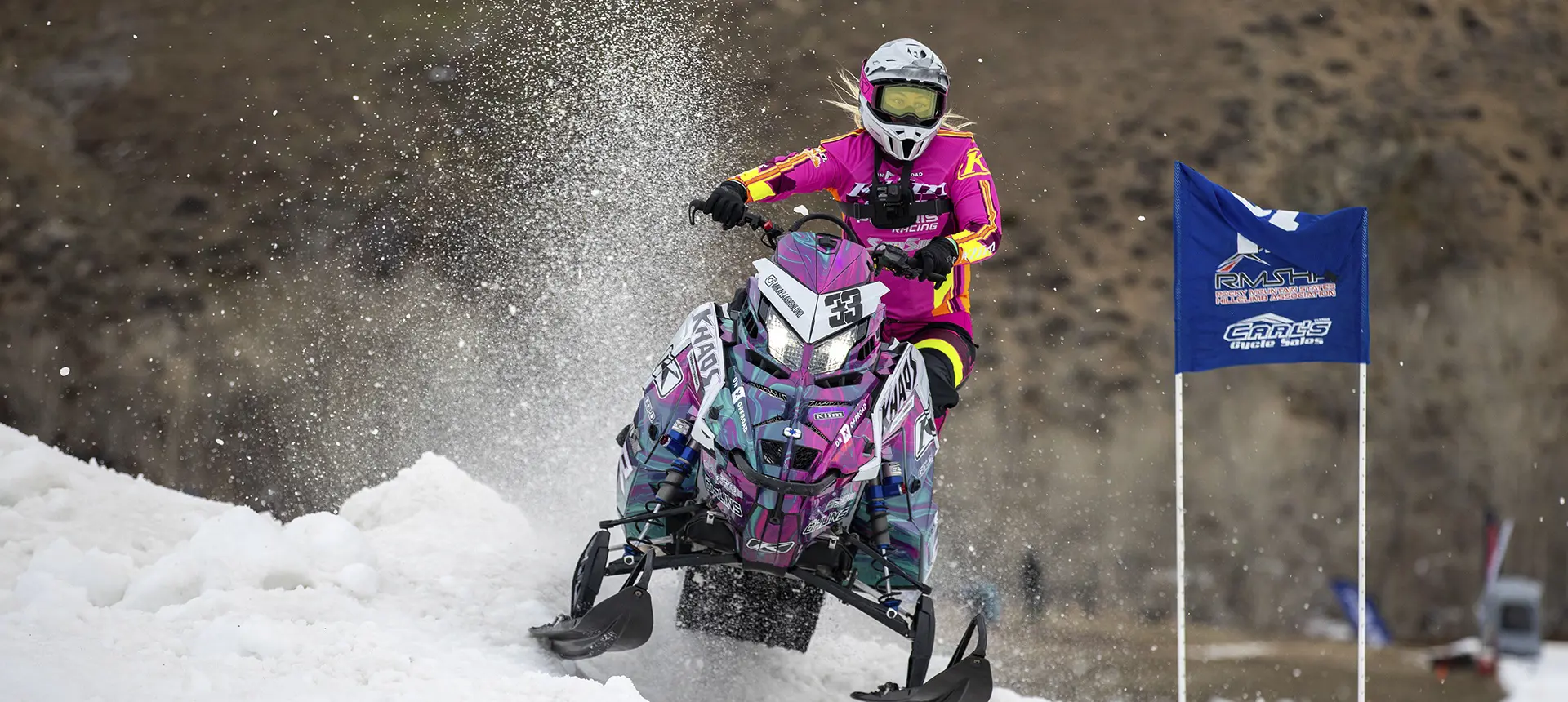 Mikaela Grönlund racing a custom Polaris RMK Khaos snowmobile through a snowy course, spraying snow near a race marker.
