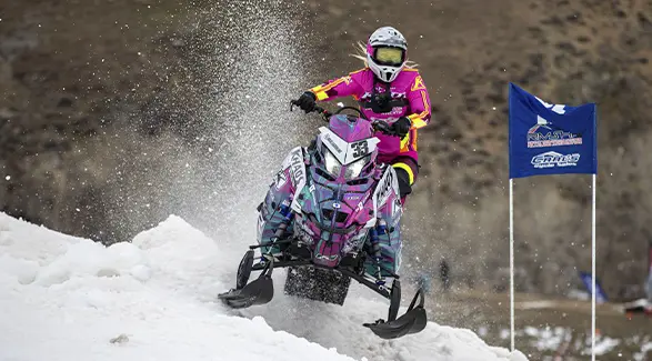 Mikaela Grönlund racing a custom Polaris RMK Khaos snowmobile through a snowy course, spraying snow near a race marker.