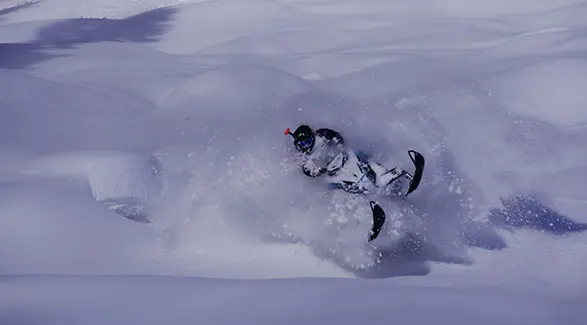 Ty Desrochers carving a Polaris snowmobile through deep mountain powder, spraying snow across the trail.