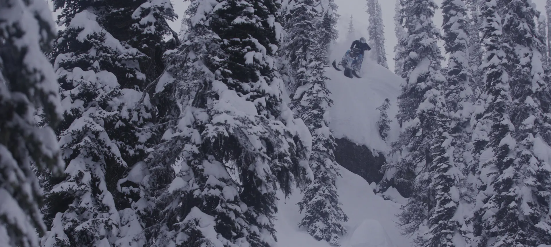 Ty Desrochers launching from a snowy cliff surrounded by snow-covered evergreen trees.