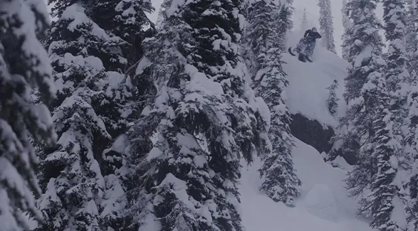 Ty Desrochers launching from a snowy cliff surrounded by snow-covered evergreen trees.