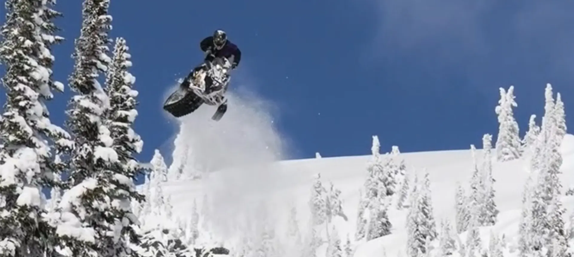 Ty Desrochers jumping a Polaris snowmobile above a snowy forest under a clear blue sky.