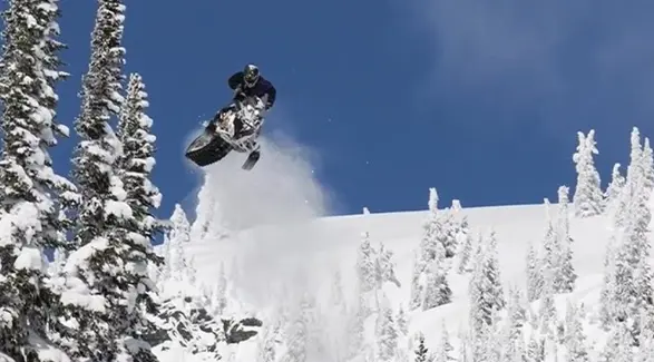 Ty Desrochers jumping a Polaris snowmobile above a snowy forest under a clear blue sky.