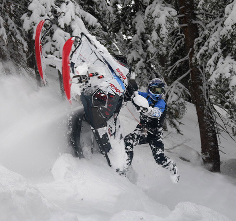 Chris Burandt carving a Polaris RMK snowmobile through deep powder in a snow-covered forest.