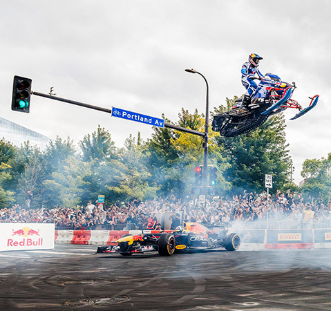 Levi LaVallee jumping a Polaris snowmobile over a Formula 1 car during a city motorsports event.