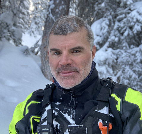 Dan Adams in black and neon yellow winter gear standing in a snow-covered evergreen forest.