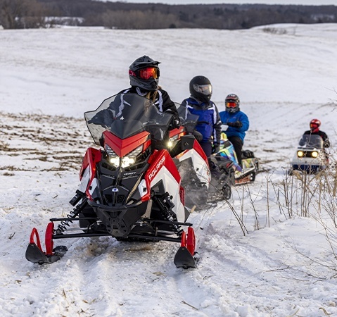 Four helmeted riders travel on Polaris snowmobiles along a snowy trail led by a red and white INDY Adventure.