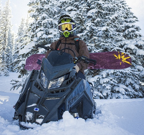 Helmeted rider on a Polaris RMK Khaos snowmobile carrying a snowboard through a snow-covered forest.
