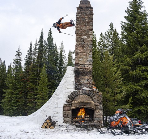Skier performing an aerial wall ride against a stone chimney surrounded by evergreen trees.