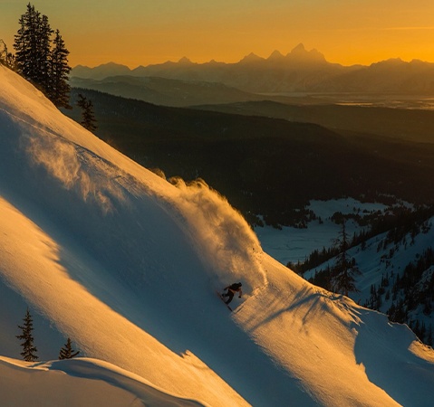 Snowboarder carving down a powder-covered mountain slope at sunset, with distant peaks on the horizon.