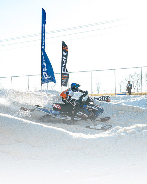 Helmeted rider racing a Polaris snowmobile through a groomed snow course lined with Polaris banners.