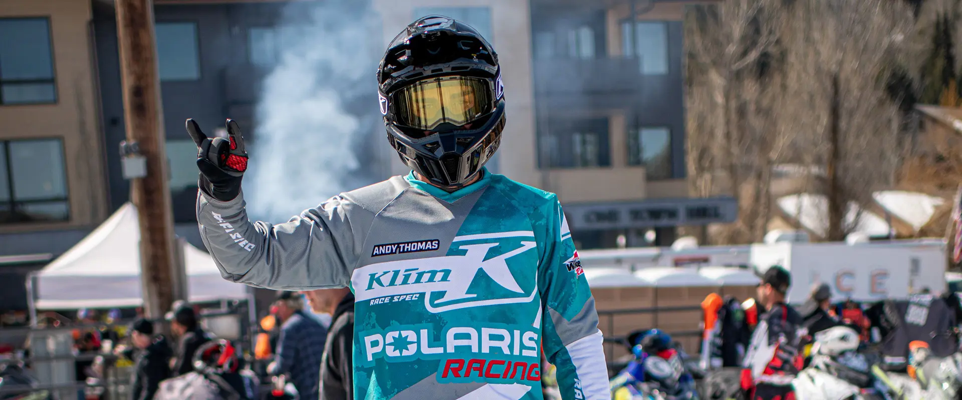 Helmeted Polaris Racing snowmobile competitor raising a hand at a crowded outdoor winter race event.