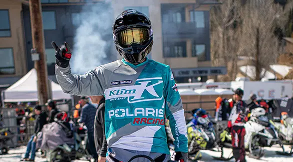 Helmeted Polaris Racing snowmobile competitor raising a hand at a crowded outdoor winter race event.