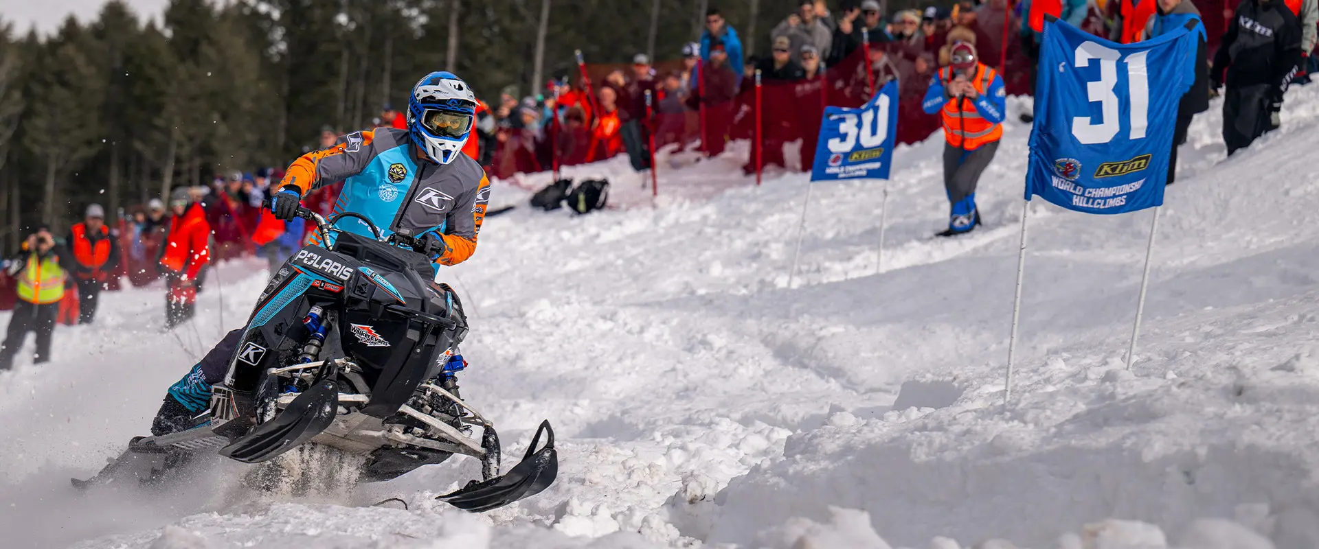 Helmeted rider racing a Polaris snowmobile through a groomed snow course lined with Polaris banners.