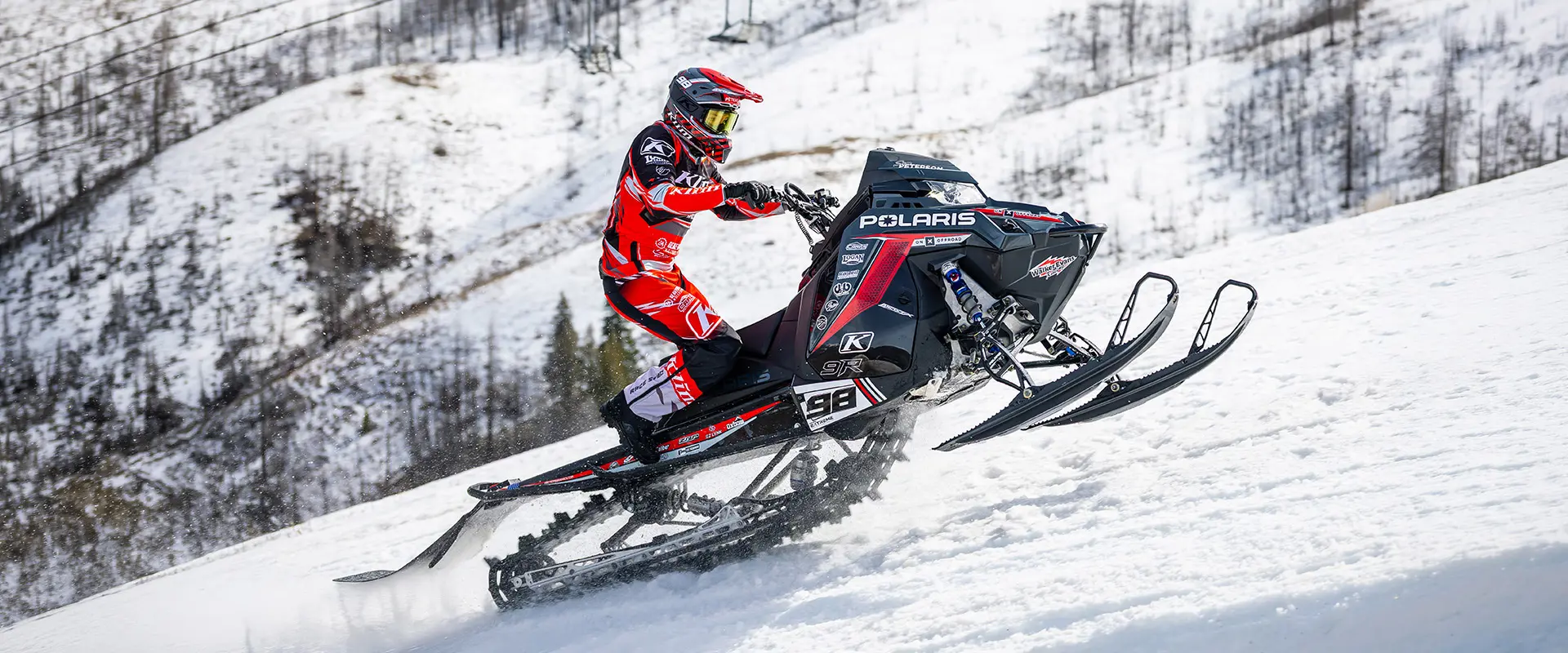 Helmeted rider racing a Polaris snowmobile uphill.