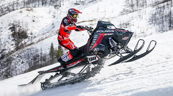 Helmeted rider racing a Polaris snowmobile uphill.