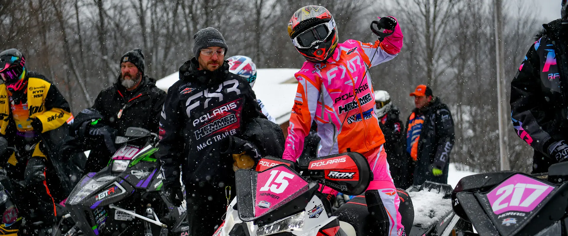 Helmeted Polaris Racing competitor beside a pink and white number 15 snowmobile at a snowy race event.