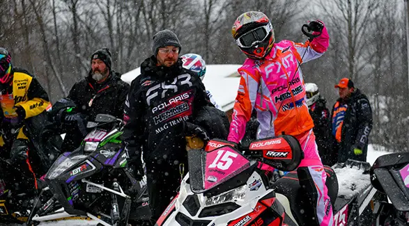 Helmeted Polaris Racing competitor beside a pink and white number 15 snowmobile at a snowy race event.