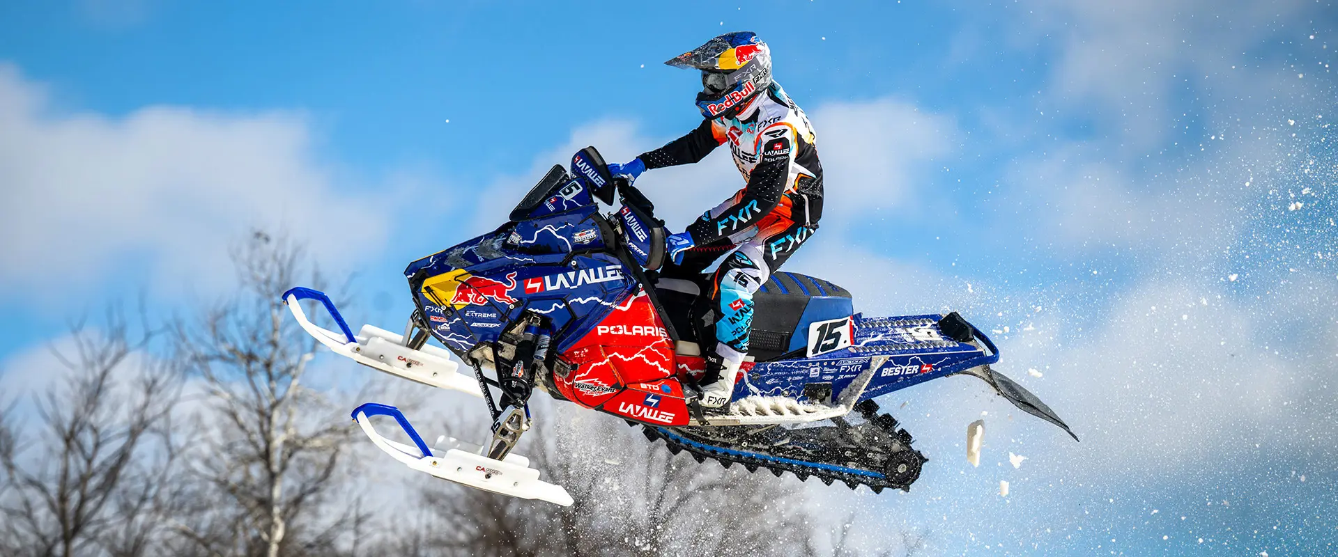 Helmeted rider jumping a blue and red number 15 Polaris race snowmobile against a clear blue sky.