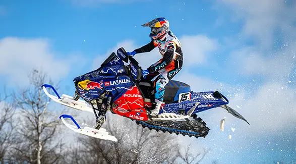 Helmeted rider jumping a blue and red number 15 Polaris race snowmobile against a clear blue sky.