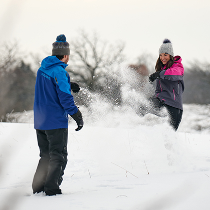 A man in a blue Polaris jacket and a woman in a grey and pink Polaris winter coat playfully kick snow at each other in an open snowy field.