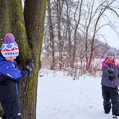 A young boy in a blue Polaris winter jacket hides behind a large tree while a girl in a pink and grey Polaris winter jacket plays in a snowy wooded area.