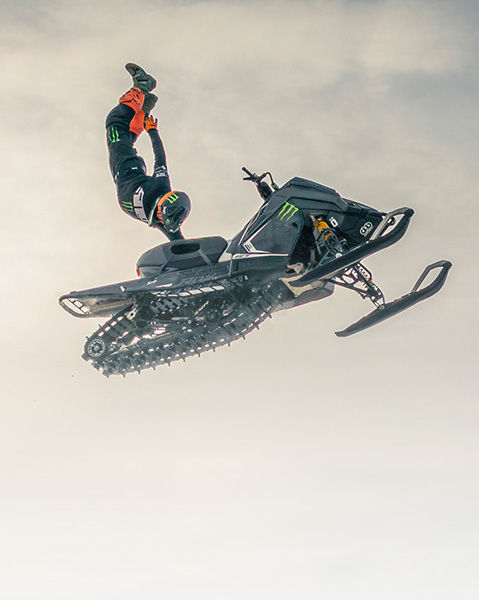 Helmeted rider performing an aerial handstand stunt above a black Polaris snowmobile.