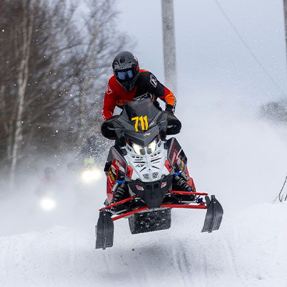 Helmeted rider racing a Polaris 600 INDY Cross Country snowmobile through deep snow on a wooded trail.