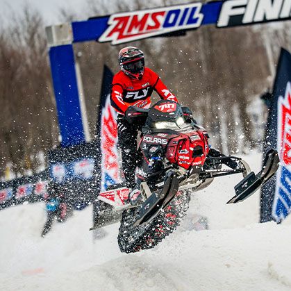 Helmeted rider launching a Polaris 600R race snowmobile over a snowy course lined with AMSOIL flags.