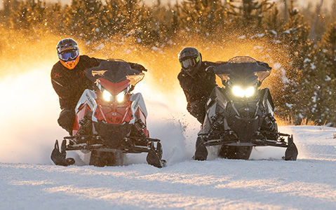 Two people on Polaris snowmobiles, one red and one black, race through deep powder at sunset, kicking up a large spray of snow.