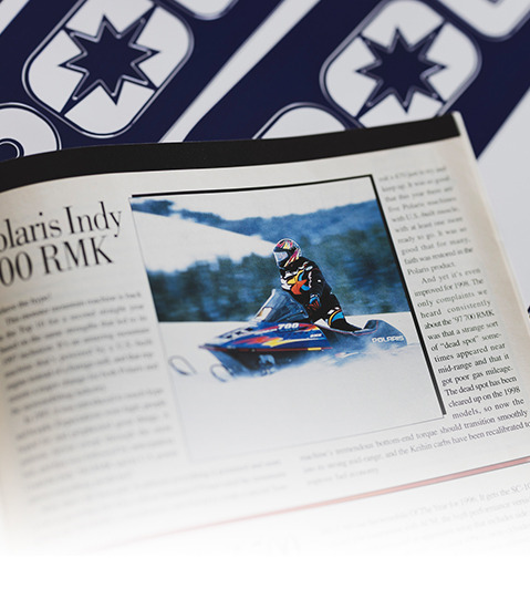 Open magazine displaying an article on Polaris Indy 700 RMK with an action photo of a rider on a blue and white snowmobile, dressed in racing gear, speeding through snowy terrain.