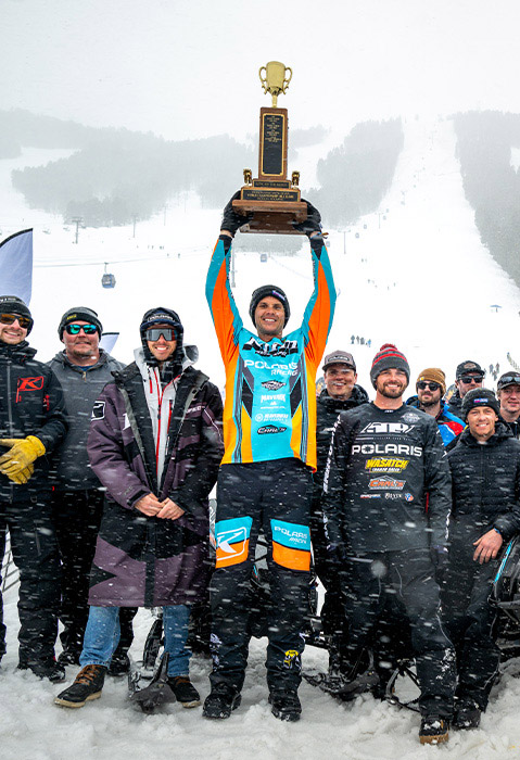 Polaris snowmobile racing team gathered in falling snow as a racer raises a championship trophy beside a race sled at a ski resort.