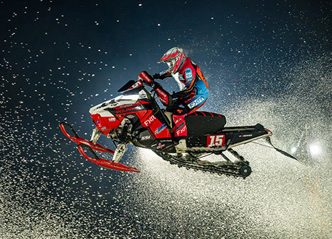 Polaris snowmobile racer airborne during a nighttime race, spraying snow beneath bright track lighting.