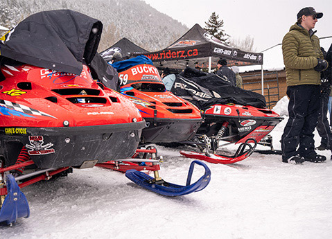 Three vintage Polaris race snowmobiles lined up in the snow beside spectators and event tents.