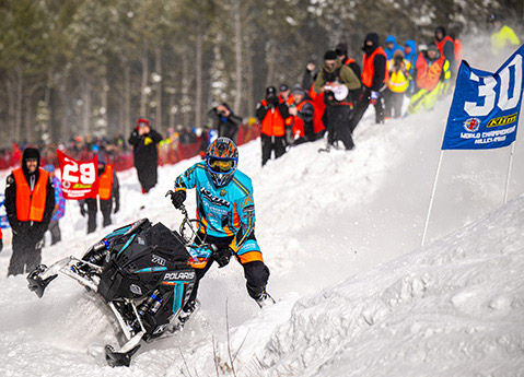 Polaris snowmobile racer navigating a steep snowbank during a championship hillclimb as spectators watch from behind the course.