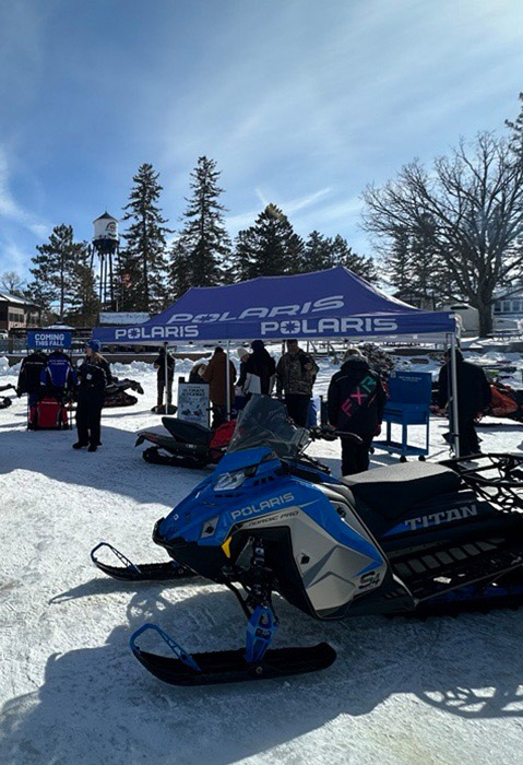 Polaris snowmobiles displayed beneath a branded tent as attendees explore a SnowCheck Sneak Peek Tour event at a snowy resort.