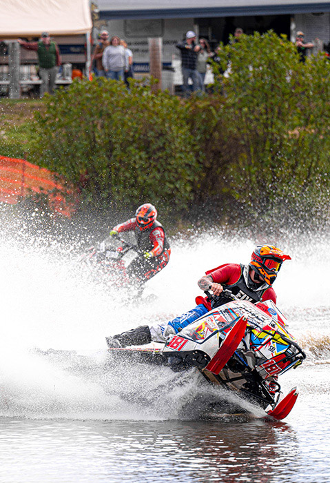 Competitive snowmobile watercross race with a rider carving through a water turn, creating a massive spray while another snowmobile follows in the background.