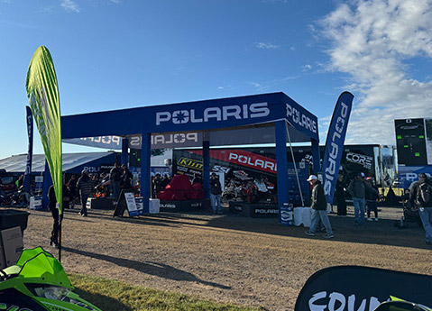 Polaris exhibitor booth at an outdoor powersports event with branded flags, display vehicles, and attendees exploring the exhibit area.