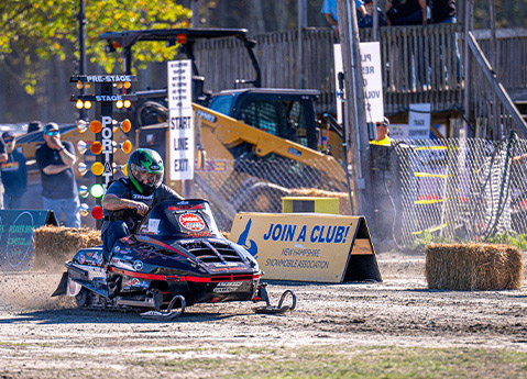 Snowmobile racer launching from the start line during a Fall Tour event on a dirt course surrounded by barriers and spectators.