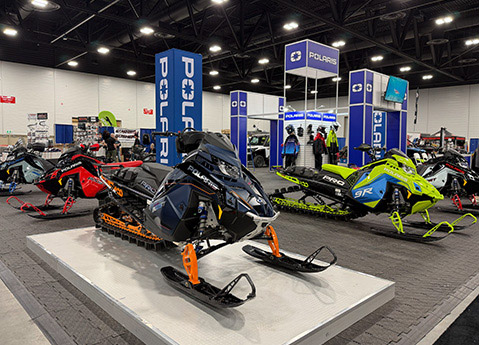 Polaris snowmobiles displayed inside an expo hall beneath branded banners, featuring mountain, trail, and crossover models.