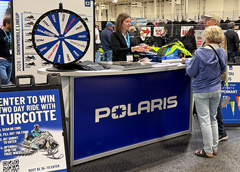 Attendees speaking with a Polaris representative at a snowmobile Fall Tour event booth featuring a prize wheel, giveaways, and Polaris sleds.
